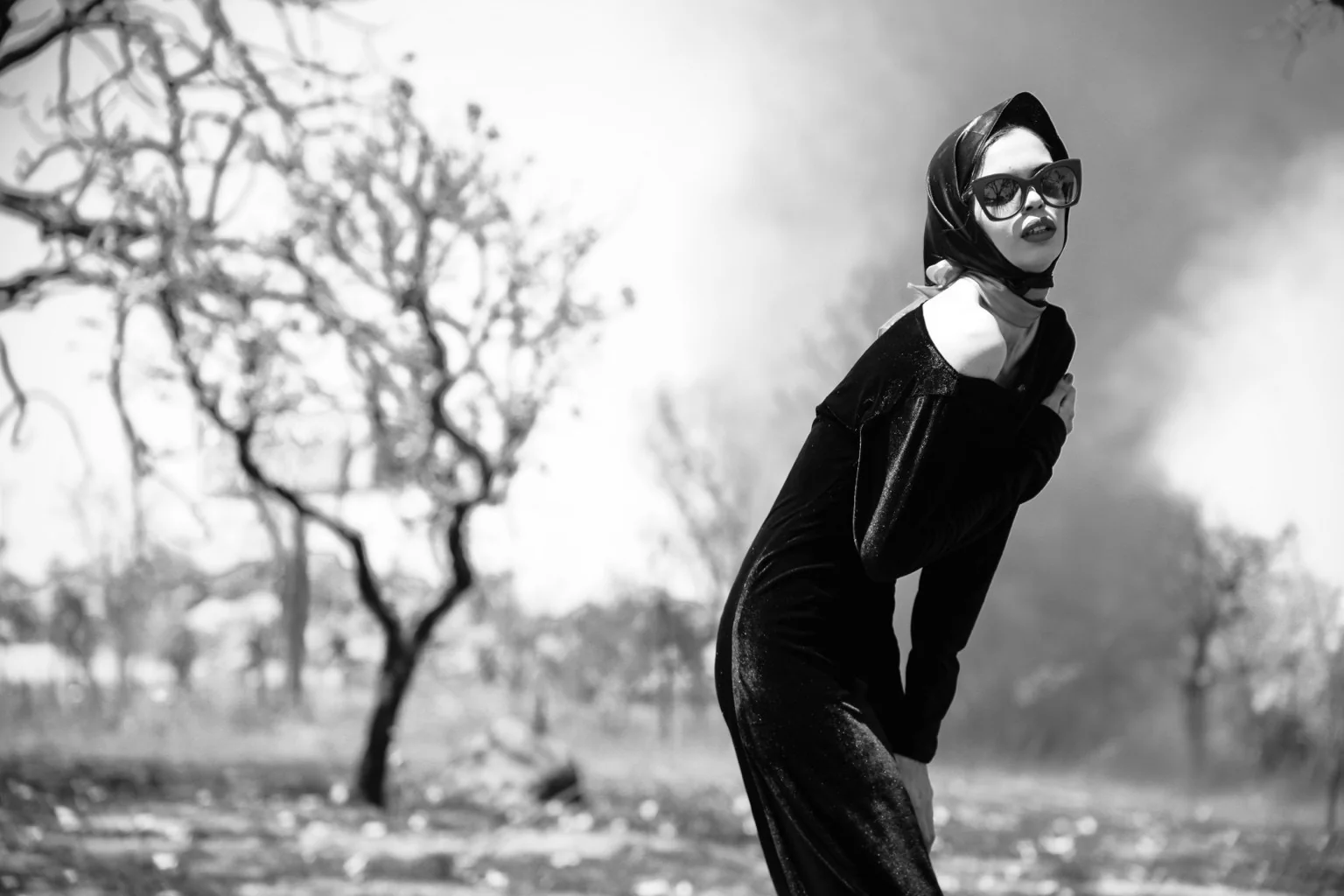 Editorial en blanco y negro: modelo con pañuelo, gafas y vestido negro entre árboles.