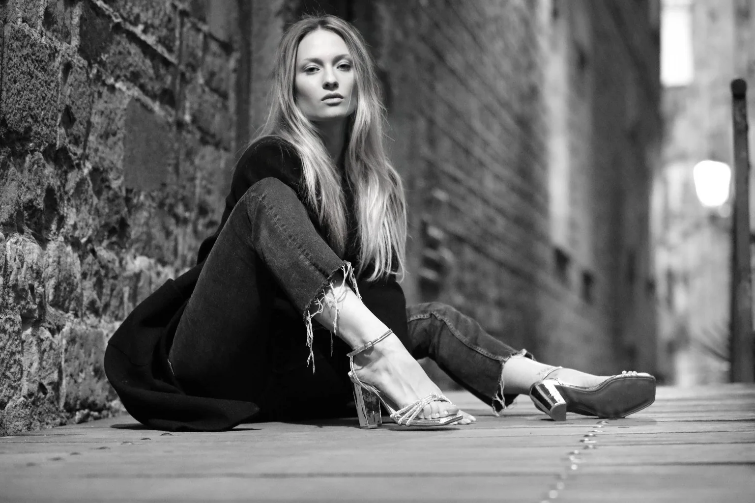 Fotografía horizontal en blanco y negro de una modelo sentada junto a un muro de piedra.