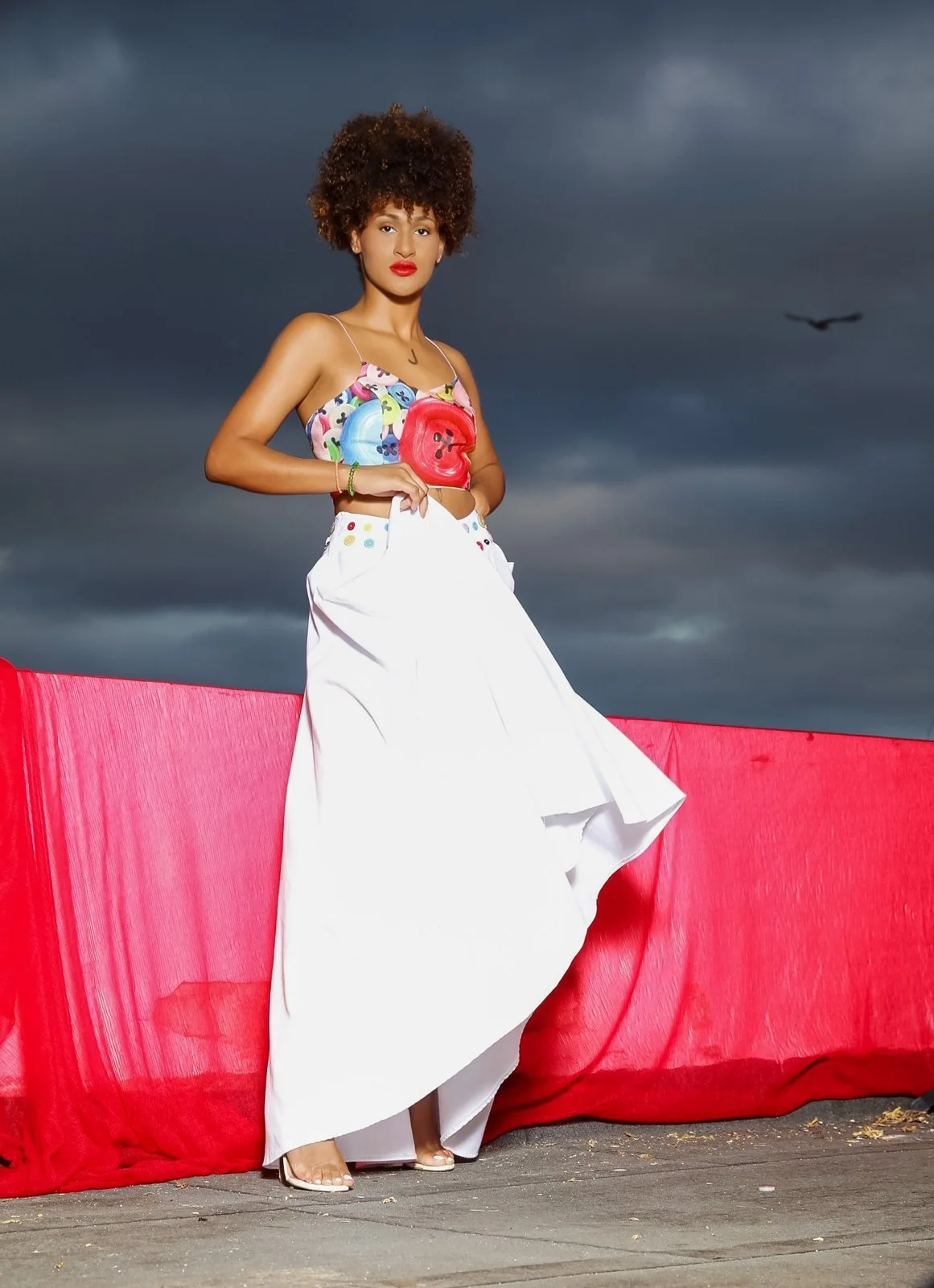 Modelo con conjunto blanco y top de colores ante tela roja y cielo oscuro.