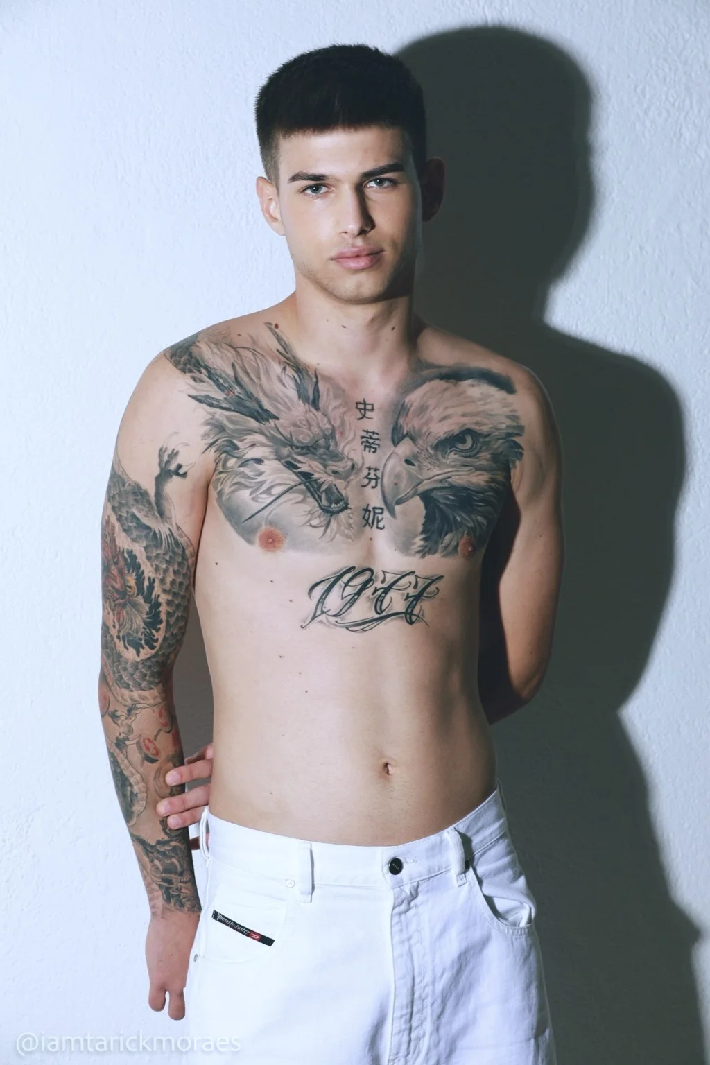 Modelo de pie con torso tatuado y sombra sobre la pared.